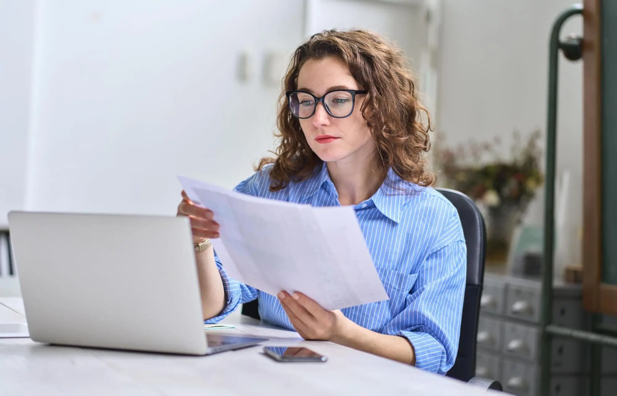 A woman with curly hair and glasses is focused on reading a document. She sits at a desk with a laptop and smartphone, in a well-lit, professional setting