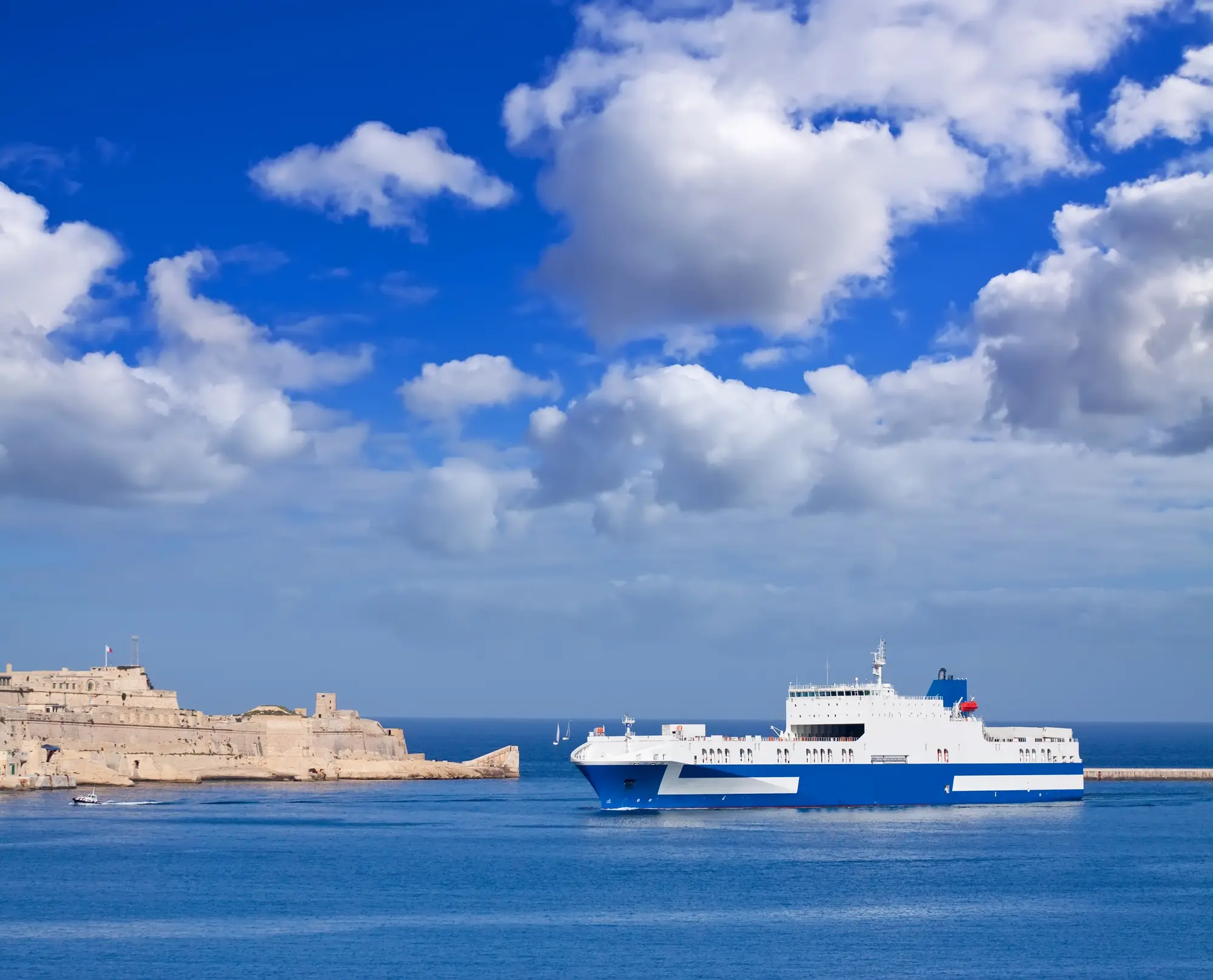 A large white and blue ferry sails through a calm blue sea under a bright sky with fluffy clouds, passing a stone fortress on a rocky coast