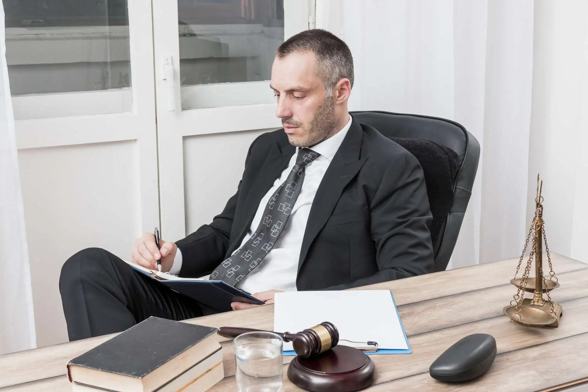 A man in a suit sits at a desk, writing on a clipboard. The desk holds a gavel, books, and scales of justice, suggesting a legal office setting
