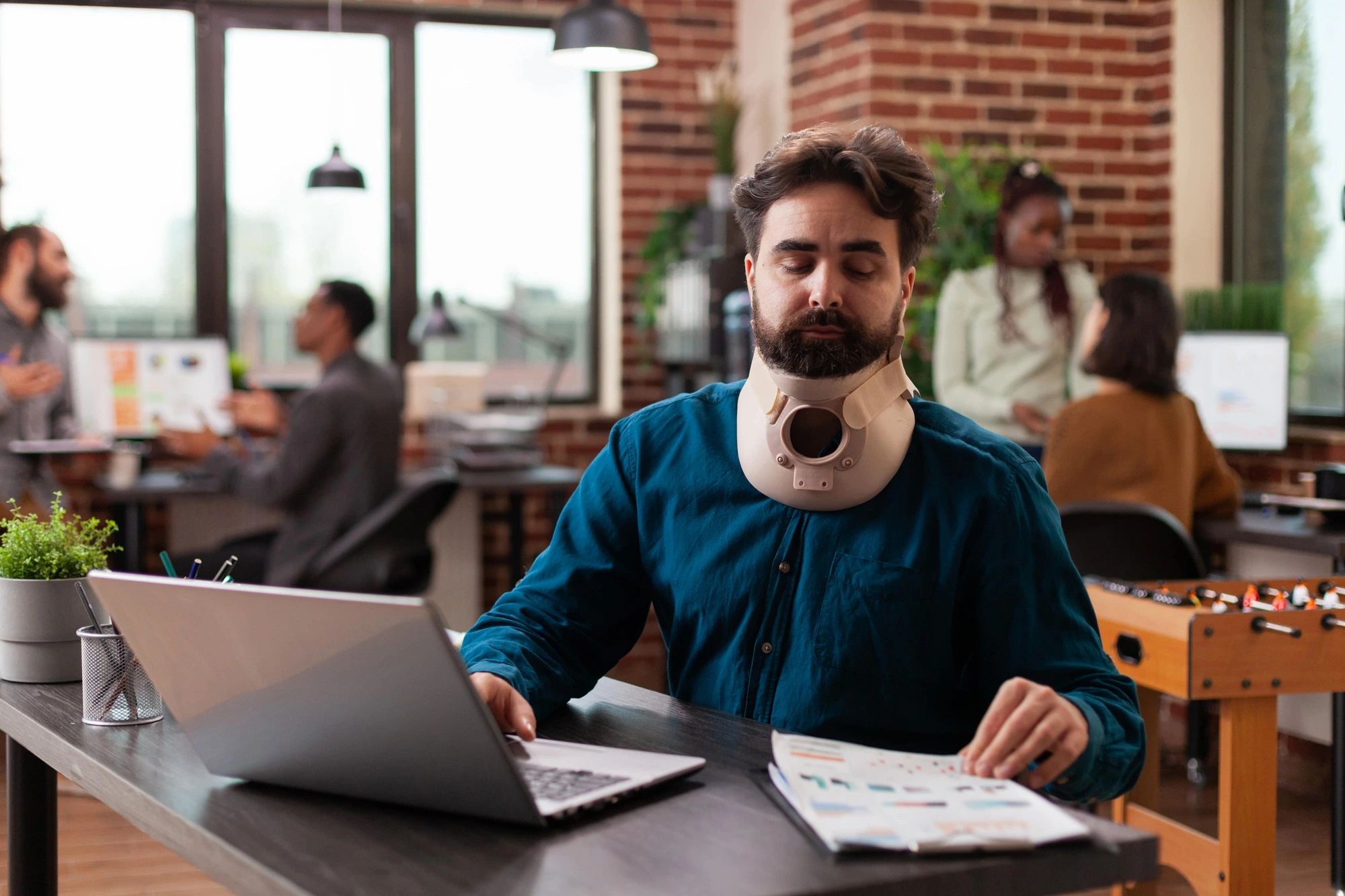 A man with a neck brace sits at a desk with a laptop and documents in a busy office. He appears focused and serious. Colleagues converse in the background