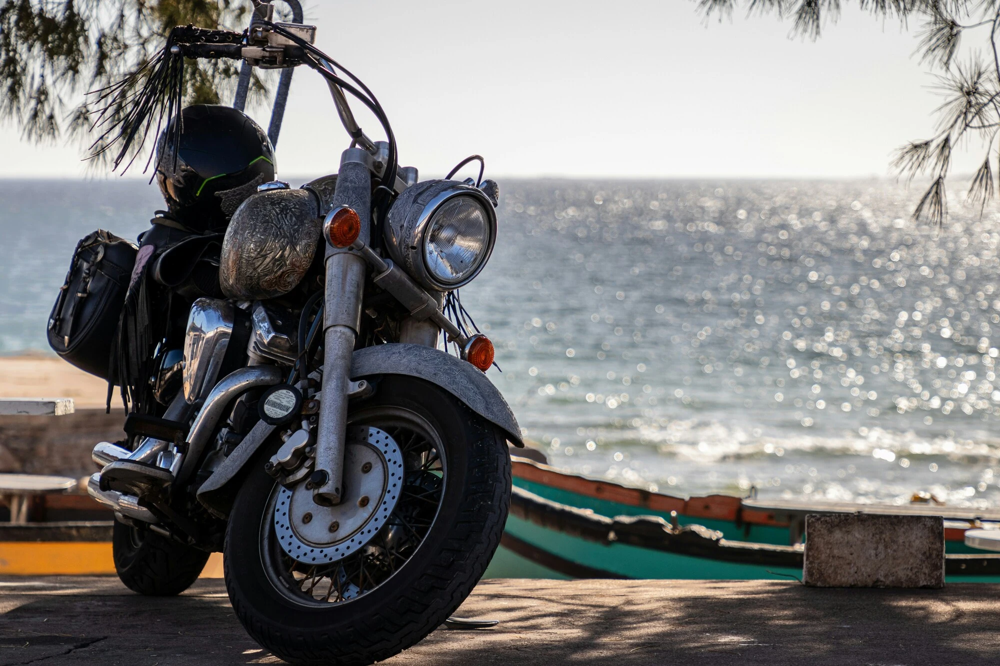 A shiny motorcycle with a helmet rests on a seaside path under a clear sky. The ocean sparkles in the background, creating a serene, adventurous vibe