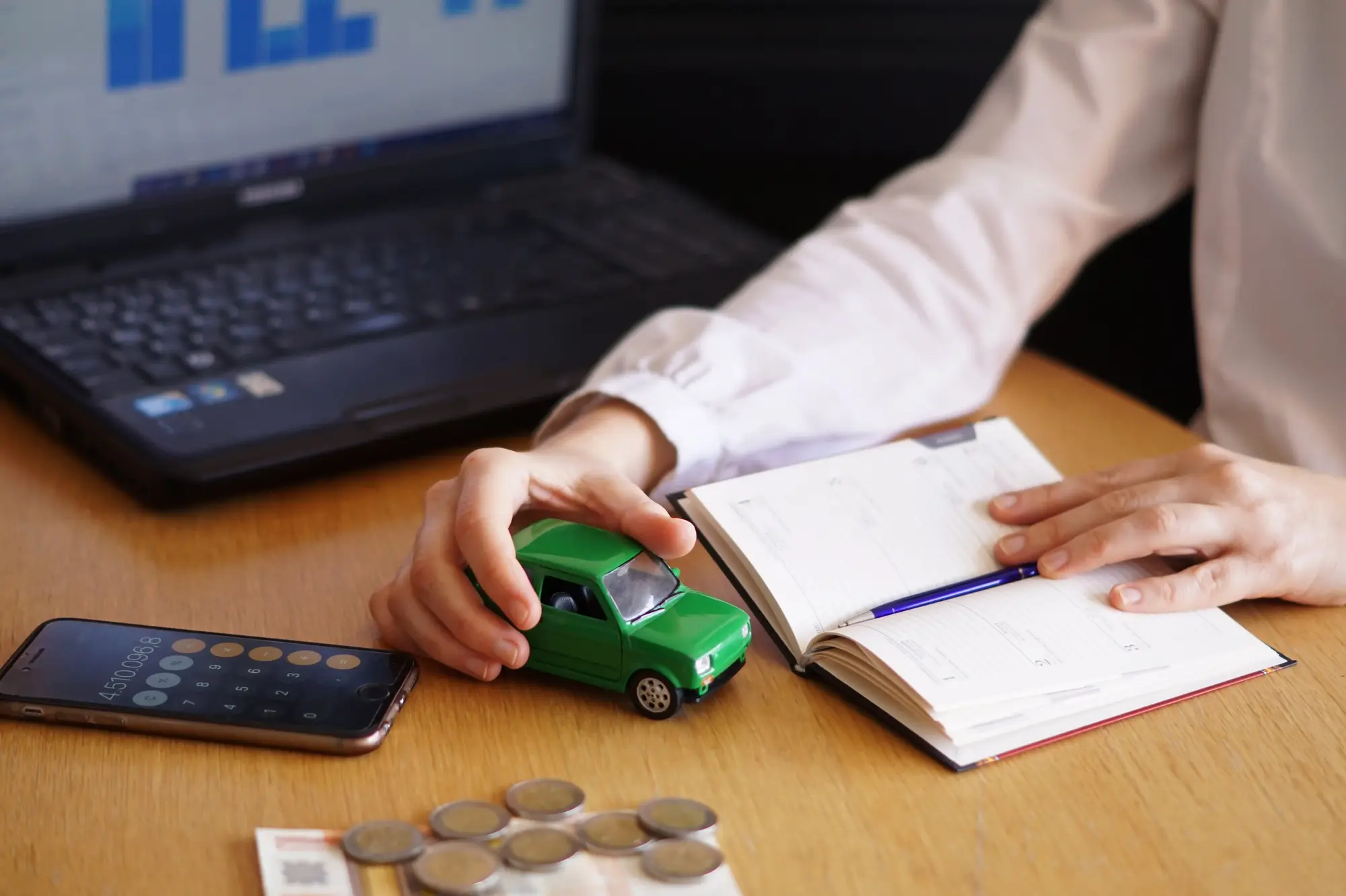 Person holding a car model with a laptop, calculator, and coins on the table, representing commercial auto insurance and business vehicle protection.