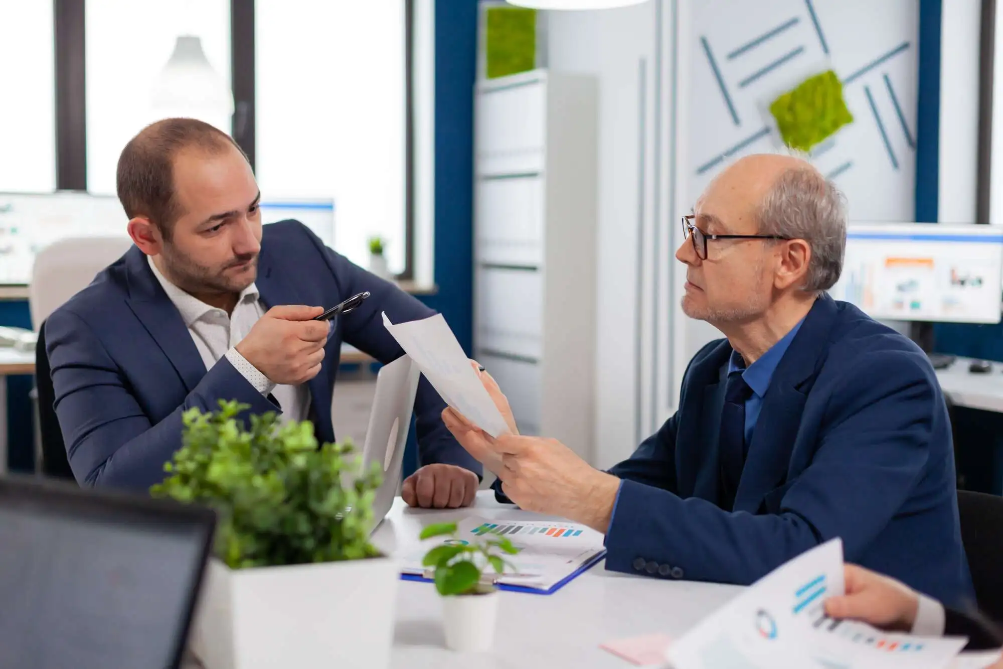 Two men in suits engaged in a serious discussion at an office table, reviewing documents. A small plant and laptop are visible, conveying a professional setting